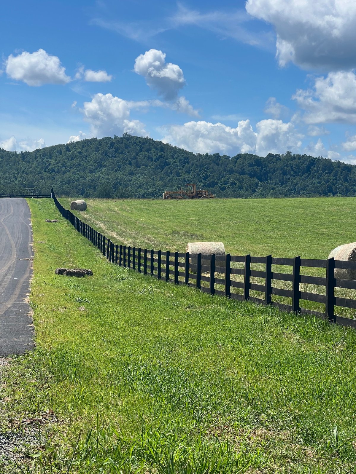 A paved road and black fence running alongside pasture toward wooded hills